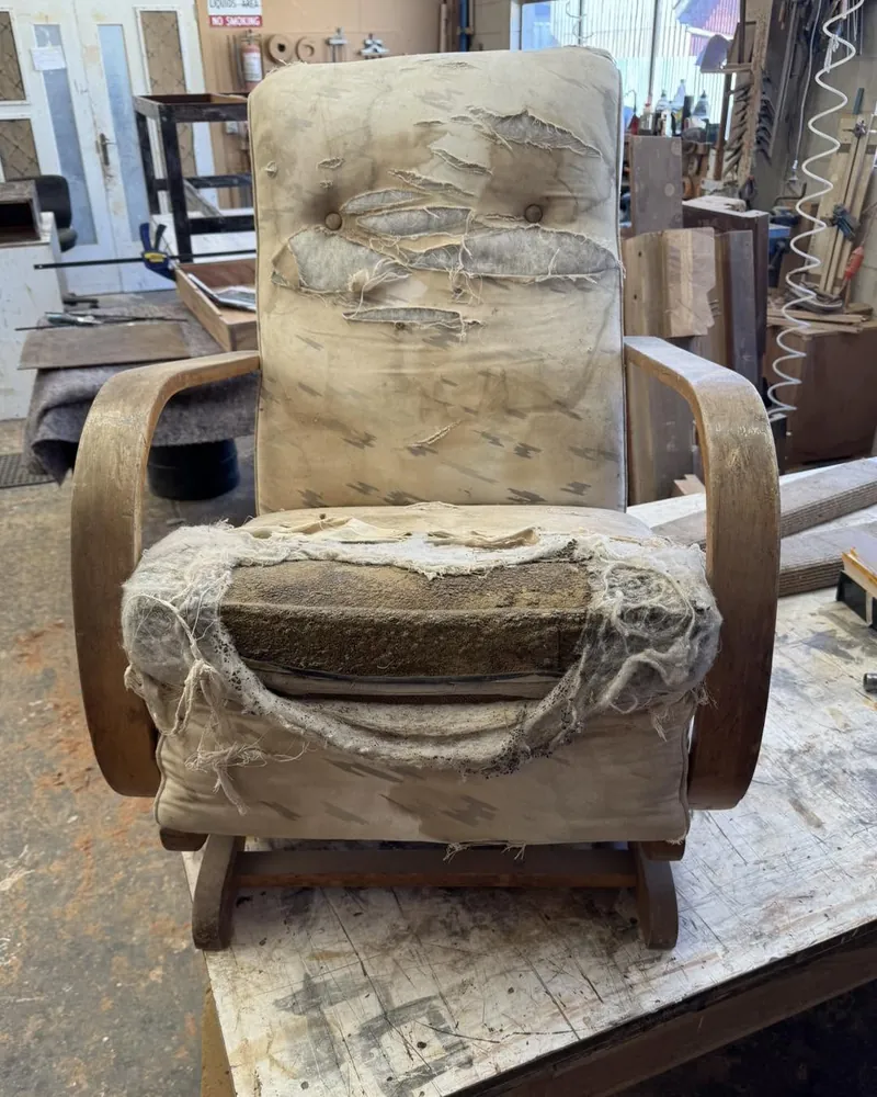 Bentwood lounge chair before restoration with torn fabric and exposed wadding on the workshop bench